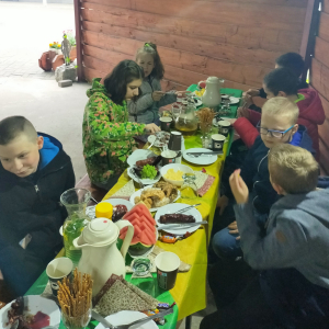 Uczniowie z klasy 5b przy stole jedzą upieczone kiełbaski oraz przygotowany poczęstunek